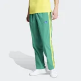 Pantalón deportivo Adidas de corte estándar, color verde con las icónicas tres rayas laterales en color amarillo. Presenta logo de la marca bordado en violeta y bolsillos laterales.