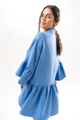 Vestido corto azul, estilo camisero, con cuello camisero, cierre frontal con botones y mangas acampanadas con volados. Presenta un diseño holgado con cortes fruncidos en el cuerpo.