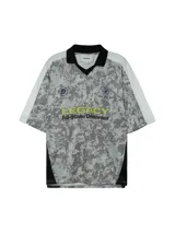 Camiseta de estilo fútbol con estampado de camuflaje gris, cuello tipo polo negro y mangas blancas. Presenta gráficos frontales con la palabra LEGACY en amarillo y detalles de escudos deportivos.