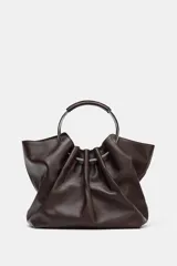 Bolso tipo saca de piel color marrón oscuro, con cuerpo fruncido y doble asa de mano rígida metálica. Incluye asa extraíble para llevar al hombro o cruzado y bolsillo interior extraíble.