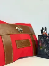 Sobre tipo cartuchera de lona roja con detalles y apliques en cuero marrón. Presenta un aplique metálico en forma de caballo y un parche de cuero con el logo de la marca Sierra Mora grabado.