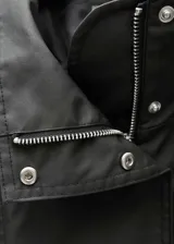 Anorak de diseño oversize en color negro, con cuello alto, cierre frontal de botones y cremallera oculta bajo tapeta. Presenta mangas largas con puños de tira abotonada, bolsillos laterales con cremallera y detalles de charreteras en los hombros.