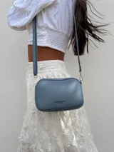 Bandolera pequeña de diseño rectangular en color beige, confeccionada en cuero sintético. Presenta una correa larga ajustable del mismo material y cierre superior con cremallera. Incluye el logo de la marca grabado en la parte frontal.