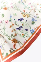 Pañuelo cuadrado de seda o material similar, color base crema, con estampado floral de flores pequeñas en tonos azules, amarillos, naranjas y rosados. Presenta un borde ancho de color terracota o marrón óxido con líneas finas de contraste.