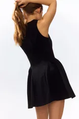 Vestido corto negro sin mangas, con escote redondo, cuerpo entallado y falda acampanada con frunces caídos en la cadera.