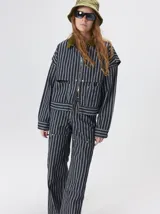 Pantalón de tiro medio y pierna ancha confeccionado en denim de algodón grueso. Presenta un diseño a rayas verticales en tonos azul y celeste, con costuras en contraste color rojo, bolsillos laterales tipo cargo y remaches metálicos.