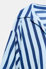 Camisa de rayas verticales azules y celestes, con cuello solapa, escote en V, cierre frontal con botones y mangas largas abullonadas con puños ajustados.