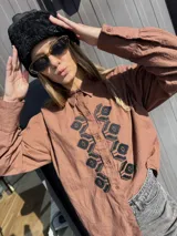Camisa de algodón con corte clásico, cuello camisero y cierre frontal con botones. Presenta un diseño bordado en punto de cruz con motivos florales en color bordó sobre el frente. Cuenta con mangas largas con puños abotonados.