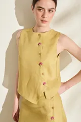 Top de lino color mostaza, sin mangas, con escote redondo y cierre frontal de botones con cartera asimétrica y detalles bordados en los ojales.