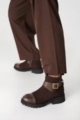 Bota corta de cuero color chocolate, con puntera reforzada en cuero liso y cuerpo en gamuza. Presenta una tira decorativa con hebilla metálica dorada en el tobillo y suela de goma tipo tractor.