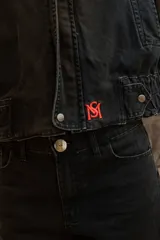 Campera oversize de jean color negro con cuello de corderito azul, cierre frontal con cremallera y solapa con botones a presión. Presenta mangas amplias y un pequeño bordado rojo en el puño.