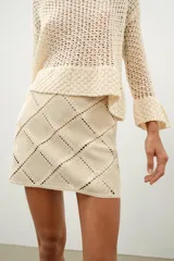 Minifalda de tiro medio confeccionada en ante color beige, decorada con aplicaciones de tejido tipo crochet blanco formando un patrón de rombos. Cuenta con cierre de cremallera oculta en la costura.
