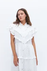 Vestido midi de lino blanco con diseño de broderie, cuello alto, cierre frontal con botones y volados decorativos en los hombros y el cuerpo.