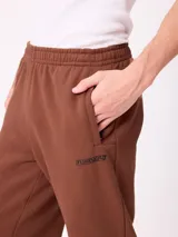 Pantalón tipo babucha de algodón y poliéster en color marrón, con cintura elástica, bolsillos laterales y logo de la marca bordado en la pierna izquierda.