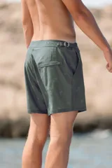 Short de baño color verde oscuro con estampado sutil de palmeras. Presenta pretina fija con hebillas metálicas laterales para ajuste, cierre frontal con botón a presión, dos bolsillos laterales y un bolsillo trasero. Incluye interior con malla hipoalergénica.