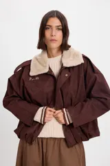 Campera bomber de gamuza color chocolate con cuello de corderito sintético desmontable en color crema. Presenta un diseño holgado con cierre frontal, hombreras decorativas y bordado sutil en el pecho.