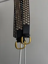 Cinturón negro de cuero ecológico con hebilla metálica dorada y doble hilera de ojales dorados a lo largo de toda la tira.