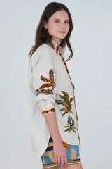 Camisa de algodón color blanco, de corte clásico con cuello camisero, manga larga y cierre frontal abotonado. Presenta un estampado tropical con palmeras y tigres, y detalles de ribetes a rayas en el cuello, la botonadura y los puños.