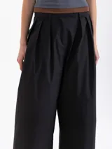 Pantalón negro de poplín asargado, corte holgado y tiro alto con pliegues frontales. Presenta un cruce en la cintura que genera un cinturón con gancho.