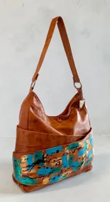 Cartera tipo hobo de cuero natural color marrón con arte pintado a mano en tonos turquesa y dorado en la base, de diseño envolvente y caída suave. Cuenta con correa de hombro y detalle de dije metálico colgante.