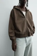 Sudadera con capucha y cierre de cremallera frontal, color marrón oscuro, de corte holgado (boxy fit) y manga larga. Presenta bolsillos frontales de vivo y acabados en rib en puños y bajo.