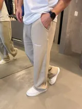 Pantalón deportivo de corte baggy y estilo oversize, color beige. Presenta costuras verticales decorativas en el frente, bolsillos laterales y un pequeño logo bordado en la pierna derecha.