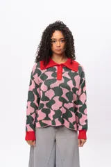 Buzo de tejido de punto con diseño estampado geométrico en tonos verde oscuro y rosado. Presenta cuello tipo polo en color rojo, cierre frontal con botones y puños acanalados en color rojo.