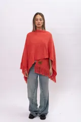 Ruana de tejido de punto color coral con flecos en el ruedo y etiqueta de cuero con logo grabado en la parte frontal.