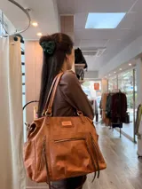 Bolso maternal de cuero marrón con doble asa de hombro, correa ajustable, múltiples bolsillos frontales y laterales con cierre, y detalles de tiras trenzadas.