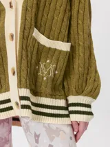 Cardigan de punto trenzado en color negro con detalles en color crema en los bolsillos, puños y ruedo. Presenta un diseño oversize, cierre frontal con botones y bordado decorativo en uno de los bolsillos delanteros.