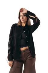 Camisa manga larga de gamuza ovina negra, con cuello clásico y cierre frontal de broches a presión. Presenta detalles de aberturas (cutouts) en las mangas entrelazadas con pasamanería de cuero en tono chocolate.