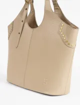 Cartera tipo shopper color beige, con diseño de costura central vertical y detalles de tachas metálicas doradas en los laterales superiores. Cuenta con correas ajustables mediante hebillas metálicas y logo grabado en la parte inferior.