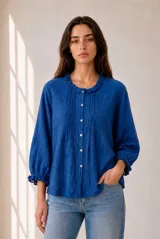Camisa azul de manga larga con textura y sutil patrón geométrico, cuello redondo con volados fruncidos y cierre frontal con botones claros. Las mangas presentan puños elásticos con terminación de volados.