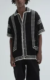 Camisa de manga corta tejida en punto abierto tipo crochet, con calce oversize y cuello solapa. Presenta un diseño bicolor en negro y blanco con botones frontales.