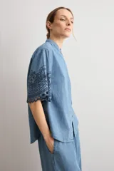 Camisa de denim celeste con cuello mao, botonadura frontal oculta y mangas cortas holgadas que presentan un delicado bordado floral oscuro y dobladillo calado.