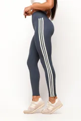 Calza deportiva de tiro alto en color azul grisáceo, con diseño de tres líneas verticales en color crema en los laterales. Confeccionada en tejido técnico de alta compresión, presenta un corte sin costura frontal para mayor comodidad.