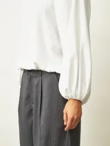 Blusa de color blanco hueso con cuello redondo, mangas largas abullonadas y puños elásticos. Presenta un diseño con frunces en los hombros y ruedo elástico que le otorga una caída holgada.
