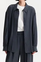 Camisa de manga larga color gris oscuro o azul marino, con cuello clásico y calce holgado. Presenta un frente con doble capa de tela que simula un canesú extendido y botones frontales.