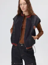 Chaleco de corte cuadrado con diseño híbrido que combina paneles de nylon acolchado y tejido de corderito. Presenta cierre frontal metálico, cuello alto y cordón ajustable en el ruedo. Incluye un parche decorativo en el pecho.