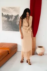 Vestido largo color beige, de silueta ajustada, con escote en V profundo, sin mangas, hombros marcados y lazo en la cintura. La falda recta cuenta con terminación de flecos en el ruedo.