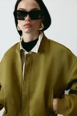 Campera bomber de tejido grueso color verde militar, con cuello corto acanalado, cierre frontal y bolsillos insertados. Cuenta con hombros caídos, mangas abullonadas y puños y bajo acanalados.