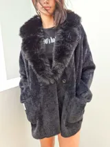 Saco negro de textura peluda con cuello de piel sintética desmontable, cierre frontal con botones y dos bolsillos delanteros.