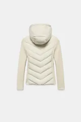 Cazadora de mujer con diseño híbrido, presenta un panel frontal acolchado con costuras en chevron y mangas y laterales en tejido liso. Cuenta con cuello alto, capucha ajustable con cordones elásticos y cierre frontal mediante cremallera.