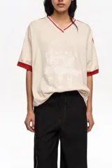 Remera oversize de color crudo con escote en V y cuello en rib rojo, mangas cortas con rib rojo y una franja roja en los hombros. Cuenta con un estampado frontal de bailarinas y detalles en relieve.