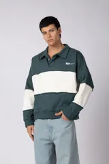 Buzo tipo polo de corte relajado y diseño color block en verde oscuro y blanco. Presenta cuello camisero con cierre de botones, mangas largas y detalles de costuras expuestas. Incluye un pequeño logo bordado en el pecho.