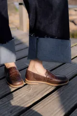 Mocasines negros de cuero con textura tipo cocodrilo en la parte superior y acabado brillante. Presentan una banda frontal clásica y una suela plana con detalle de borde en color natural.