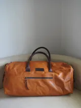 Bolso de viaje tipo weekender de cuero color tostado con asas y detalles en marrón oscuro, herrajes metálicos y bolsillo frontal con cierre.