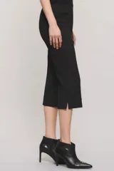 Pantalón capri negro de corte sastrero, confeccionado en tejido de punto grueso con elastano. Presenta calce slim, alforzas en el delantero, bolsillos ojal simulados, aberturas laterales en los bajos y cierre frontal con botón y broche metálico.