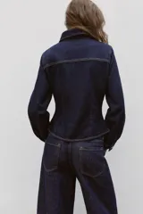 Camisa entallada de denim azul oscuro con cuello solapa, manga larga y cierre frontal con botones metálicos. Presenta un bolsillo de plastrón en el pecho y costuras a contratono.
