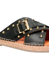 Sandalia tipo ojota con base de yute, tiras cruzadas de cuero negro con tachas doradas y hebilla metálica decorativa.